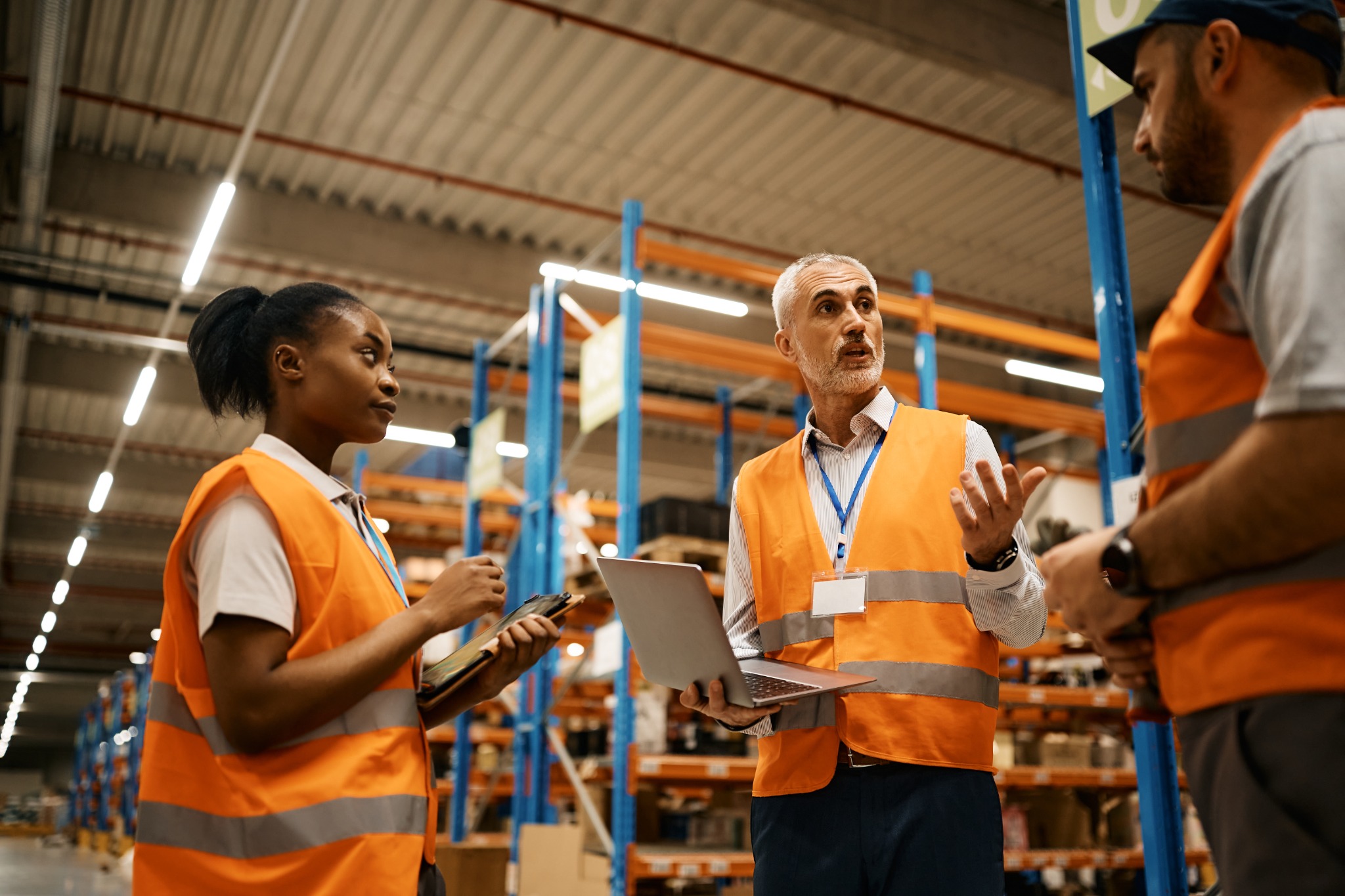 A multigenerational operations team reviewing work together on a laptop and tablet in a warehouse aisle.